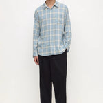 A man modeling a light blue and white plaid long-sleeve button-up shirt with black trousers, standing against a plain white studio background.