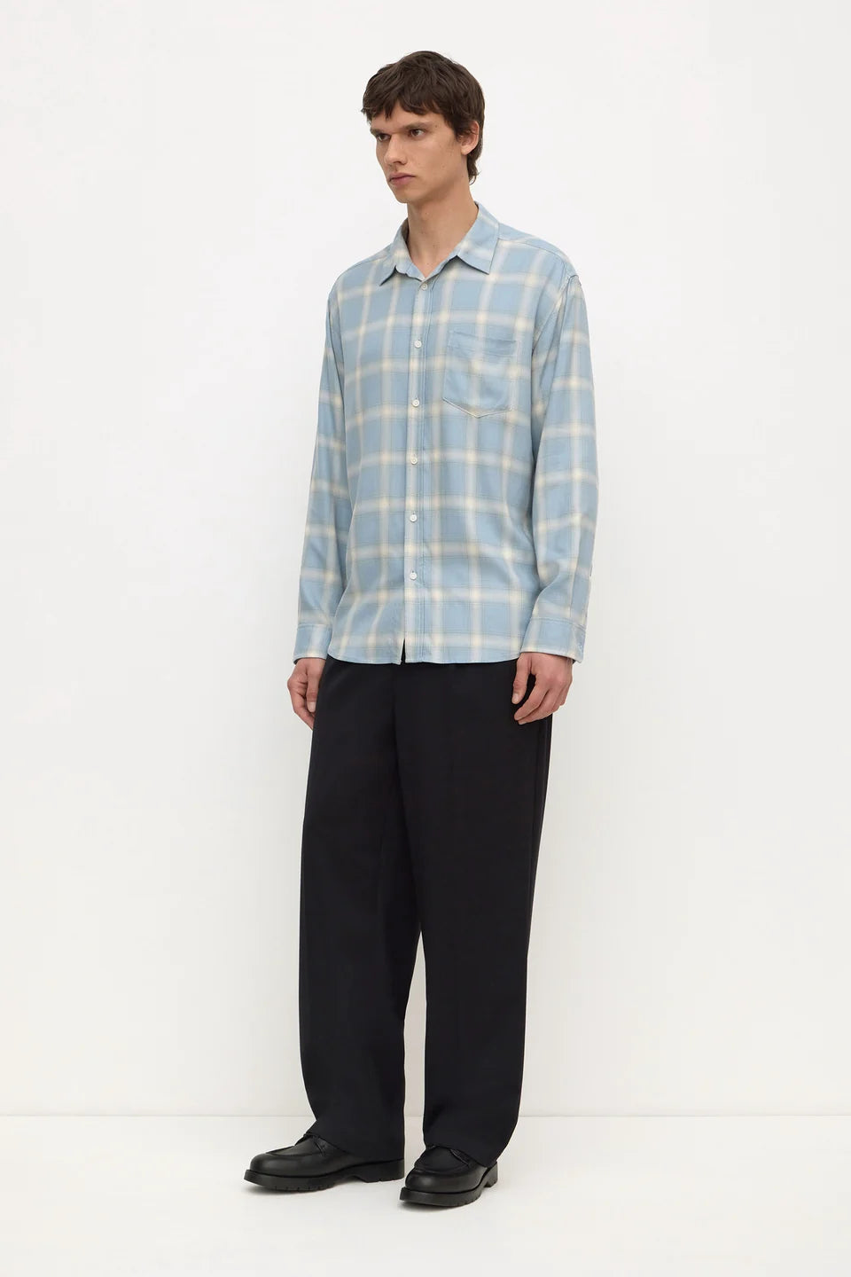 A man modeling a light blue and white plaid long-sleeve button-up shirt with black trousers, standing against a plain white studio background.