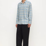 A man modeling a light blue and white plaid long-sleeve button-up shirt with black trousers, standing against a plain white studio background.