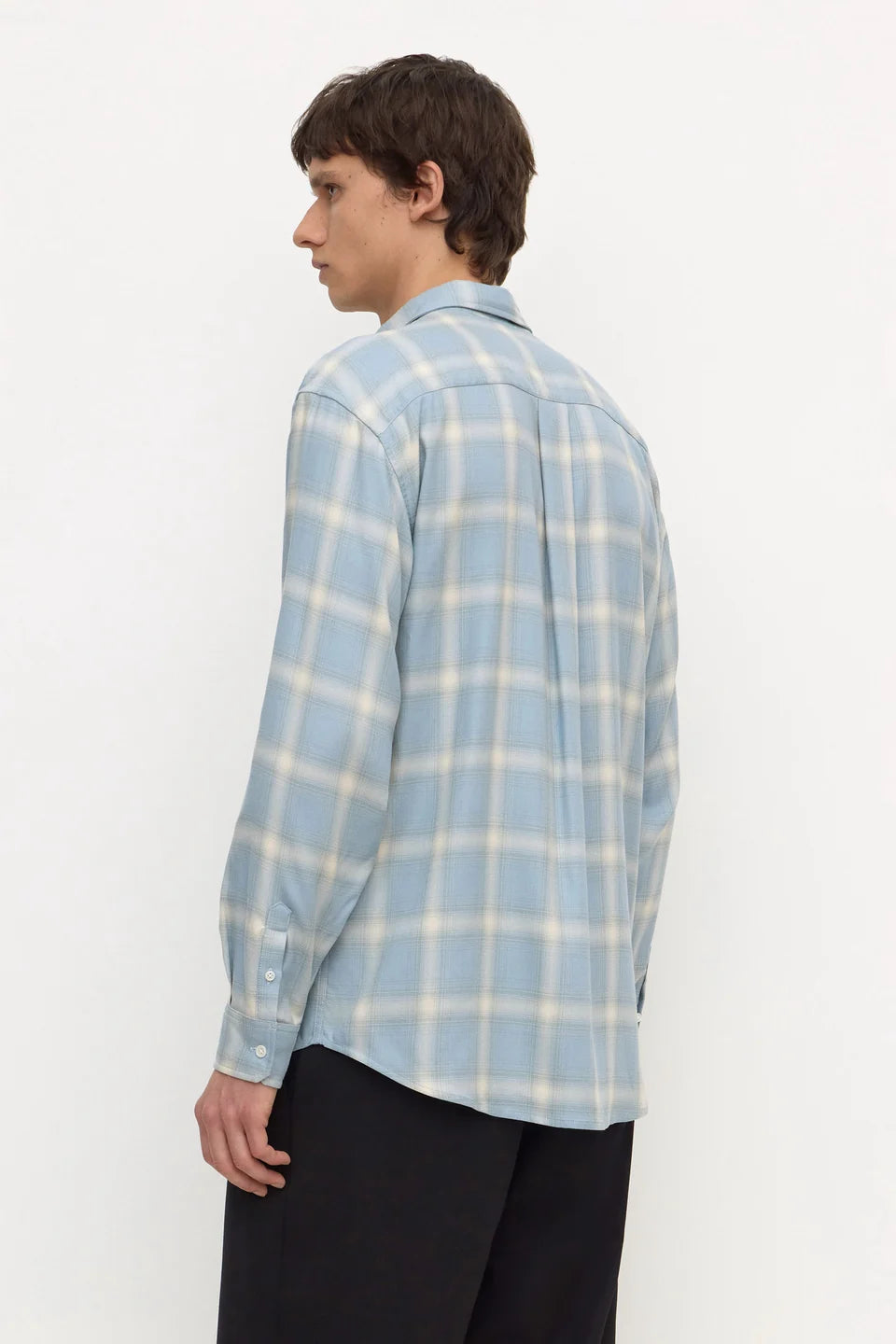 A man wearing a light blue and white plaid long-sleeve button-up shirt, shown from a side-back angle against a plain white studio background.