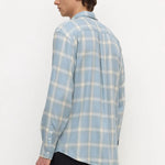 A man wearing a light blue and white plaid long-sleeve button-up shirt, shown from a side-back angle against a plain white studio background.