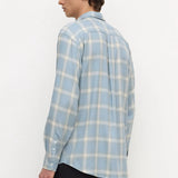 A man wearing a light blue and white plaid long-sleeve button-up shirt, shown from a side-back angle against a plain white studio background.