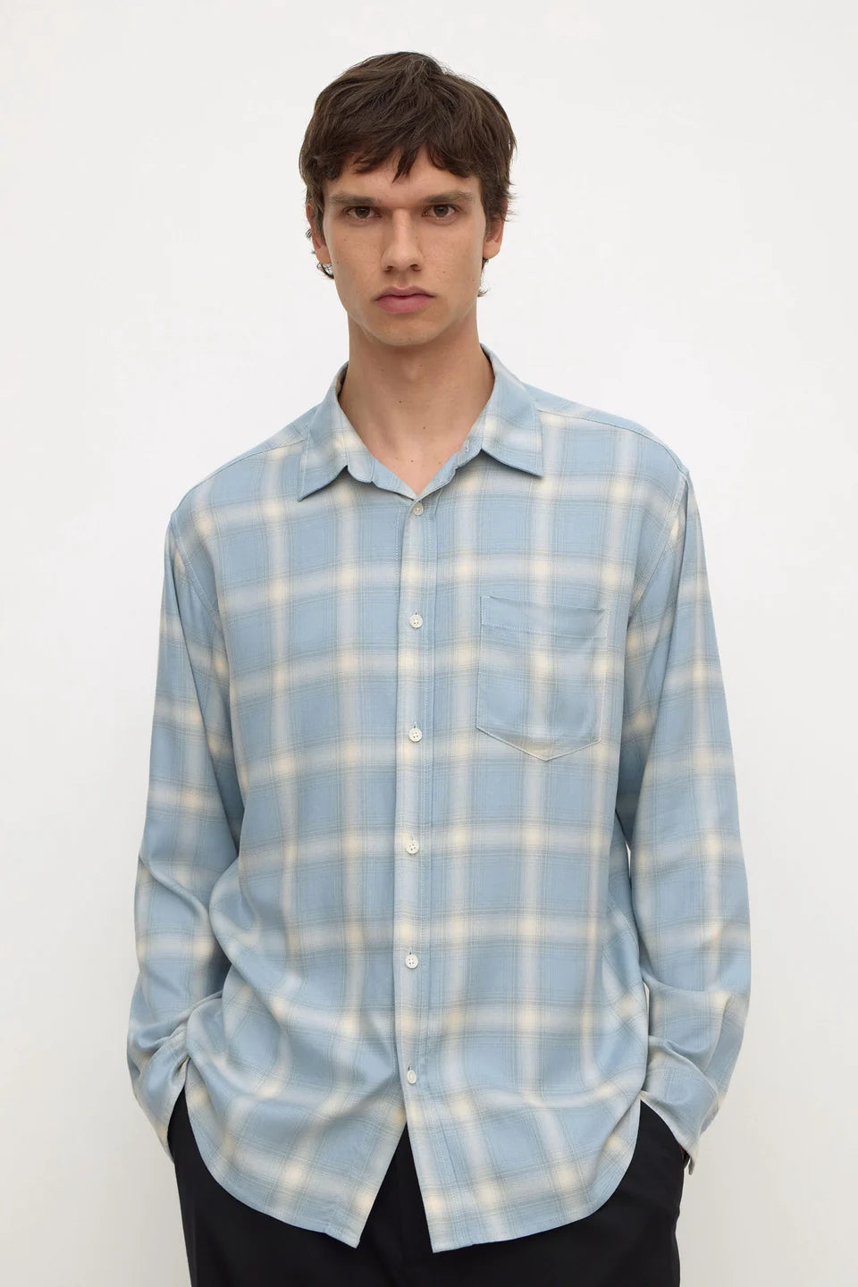 A man wearing a light blue and white plaid button-down shirt with a chest pocket, paired with black pants, standing against a plain white background.