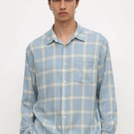A man wearing a light blue and white plaid button-down shirt with a chest pocket, paired with black pants, standing against a plain white background.