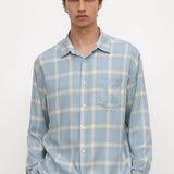 A man wearing a light blue and white plaid button-down shirt with a chest pocket, paired with black pants, standing against a plain white background.