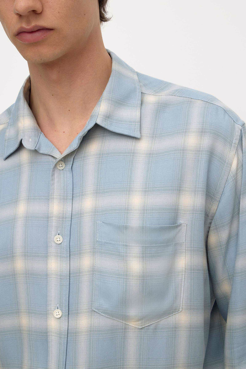 A close-up of a person wearing a light blue and white plaid button-up shirt with a chest pocket, shown against a plain light background.