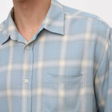 A close-up of a person wearing a light blue and white plaid button-up shirt with a chest pocket, shown against a plain light background.