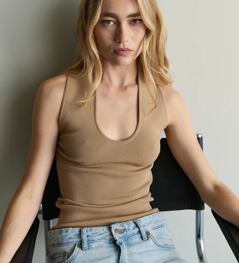 Woman wearing a beige tank top and blue jeans sitting on a chair against a plain background