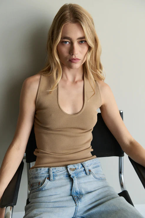 Woman wearing a beige tank top and blue jeans sitting on a chair against a plain background