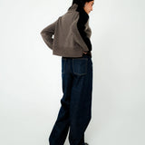A model is shown from a side-back angle wearing a cropped taupe-brown knit cardigan with black side panels, dark wide-leg jeans, and black shoes, set against a plain white studio background.