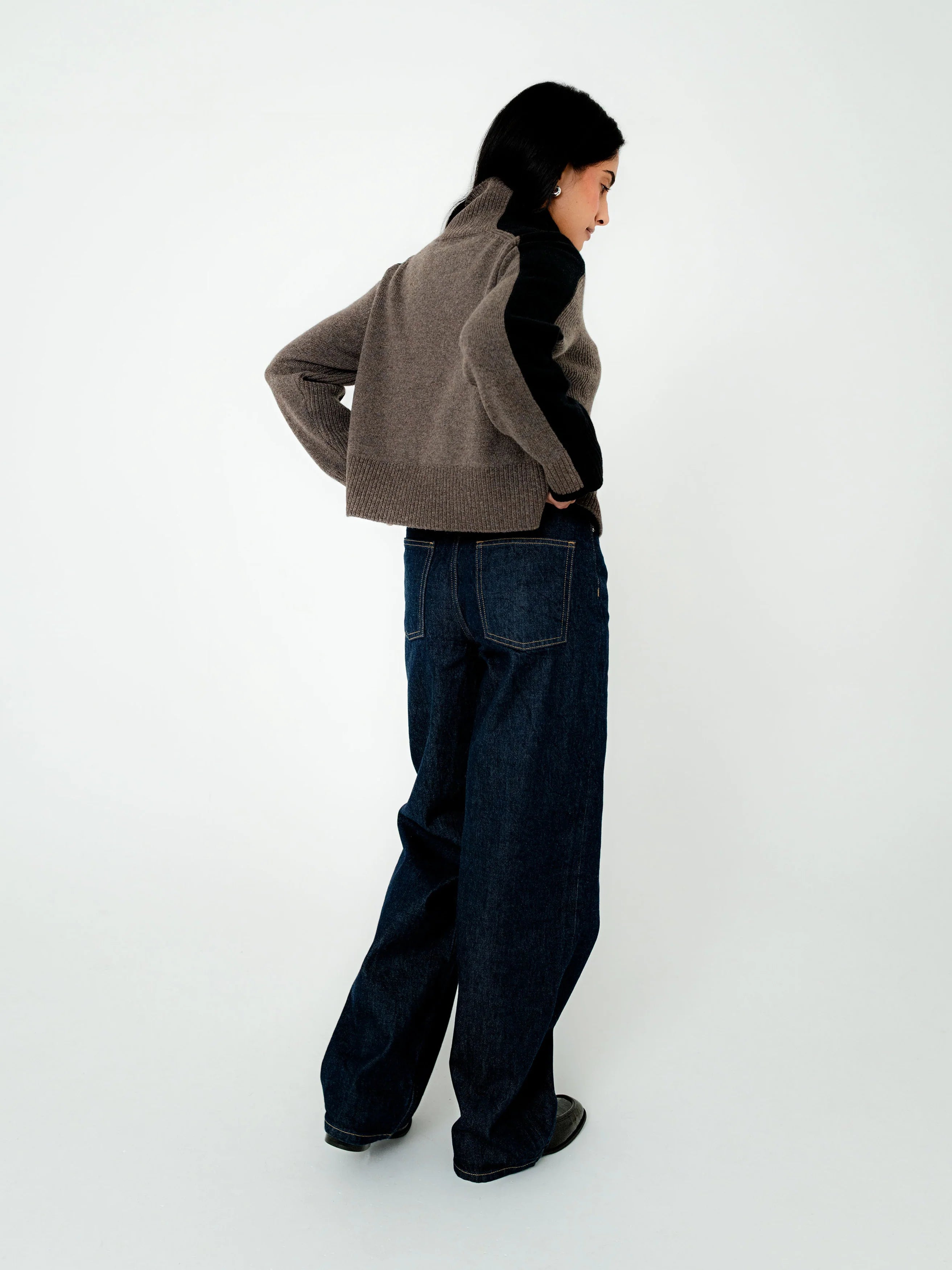 A model is shown from a side-back angle wearing a cropped taupe-brown knit cardigan with black side panels, dark wide-leg jeans, and black shoes, set against a plain white studio background.