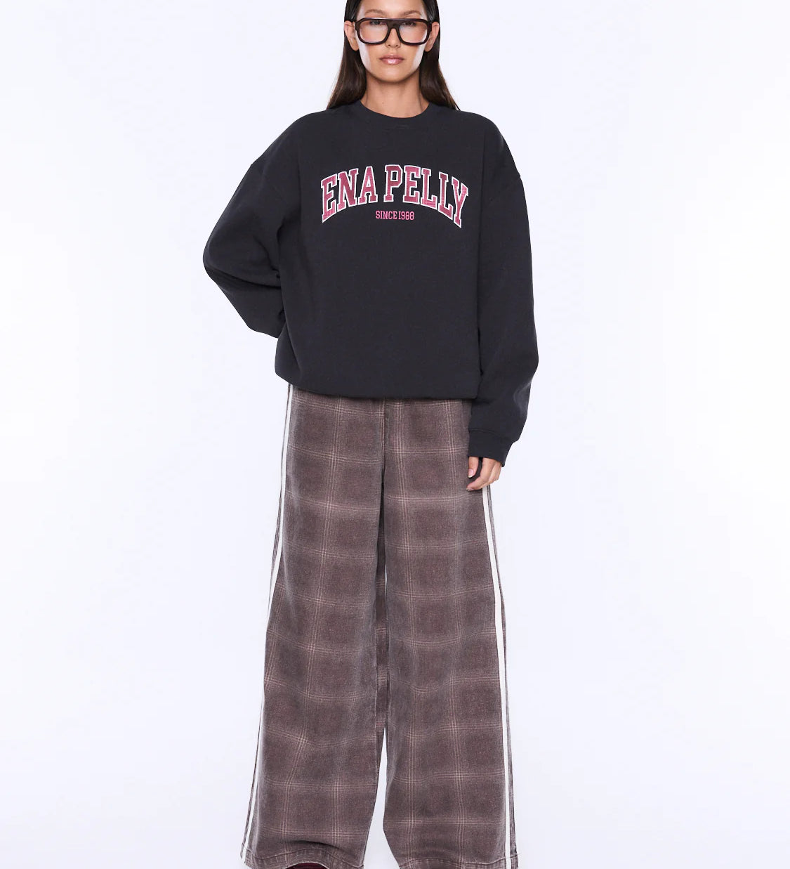 A model wearing a black crewneck sweatshirt with 'ENA PELLY' printed in pink and white on the chest, paired with wide-leg brown plaid pants featuring white side stripes, set against a plain white studio background.