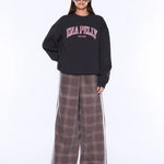 A model wearing a black crewneck sweatshirt with 'ENA PELLY' printed in pink and white on the chest, paired with wide-leg brown plaid pants featuring white side stripes, set against a plain white studio background.
