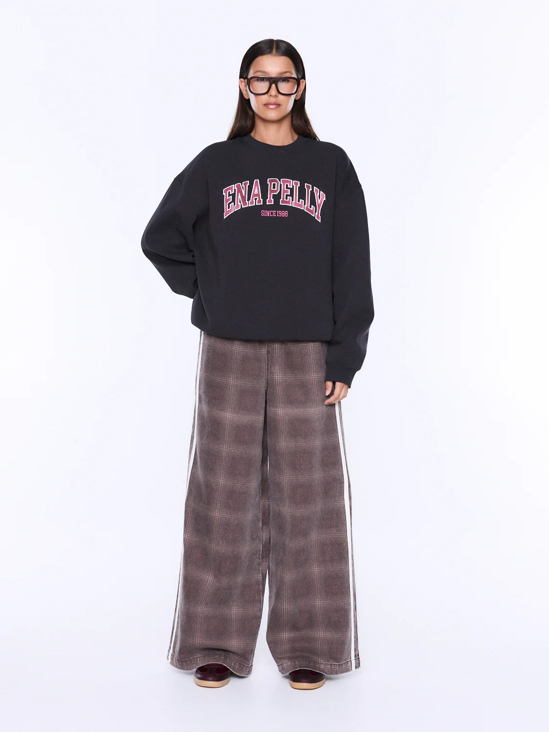 A model wearing a black crewneck sweatshirt with 'ENA PELLY' printed in pink and white on the chest, paired with wide-leg brown plaid pants featuring white side stripes, set against a plain white studio background.