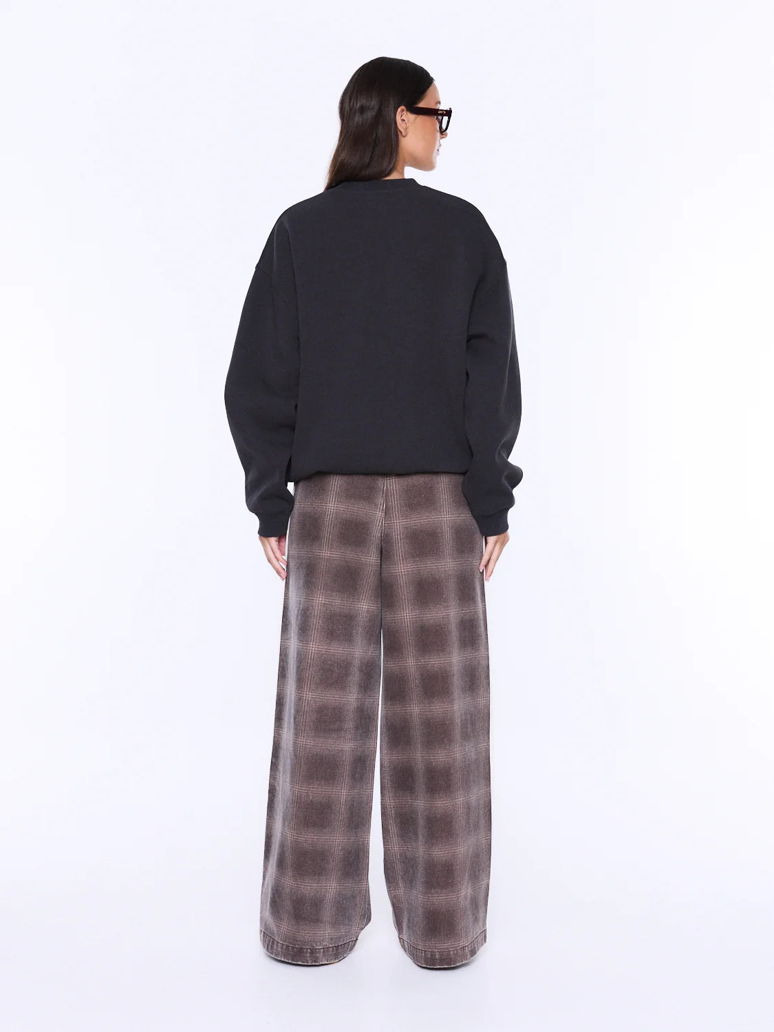 A model shown from the back wearing a black crewneck sweatshirt and wide-leg plaid trousers in brown and gray, photographed against a solid white studio background.