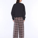 A model shown from the back wearing a black crewneck sweatshirt and wide-leg plaid trousers in brown and gray, photographed against a solid white studio background.