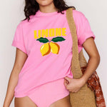 Woman wearing a pink t-shirt with 'Limone' design and sunglasses, holding a straw bag.