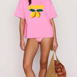 Woman wearing a pink t-shirt with lemon graphic and text, holding a straw bag on a plain background