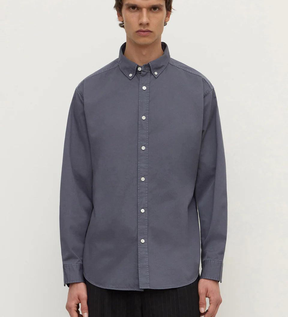 A man wearing a slate blue long-sleeve button-down shirt with a button collar, paired with dark trousers, standing against a plain light gray background.