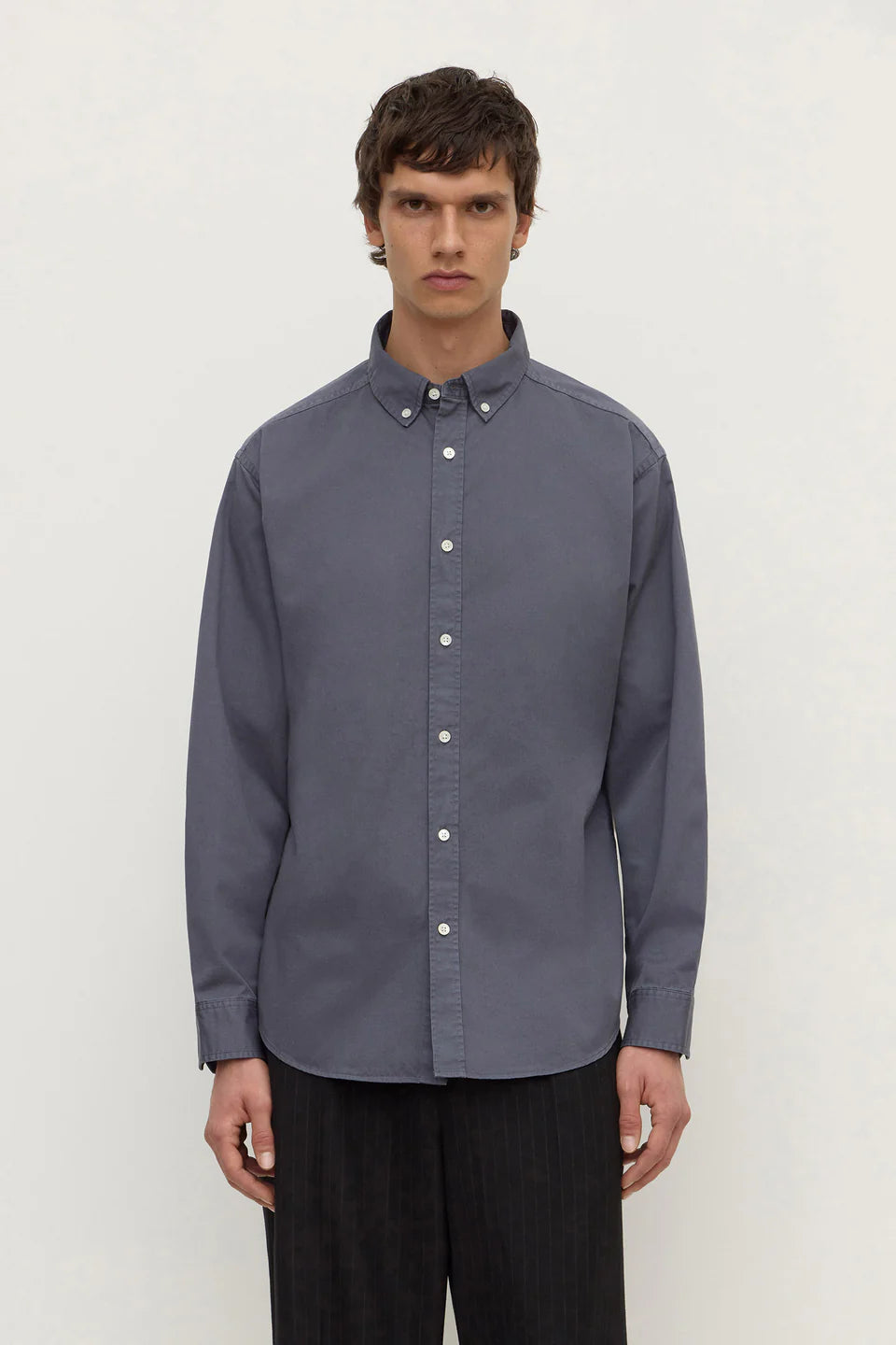 A man wearing a slate blue long-sleeve button-down shirt with a button collar, paired with dark trousers, standing against a plain light gray background.