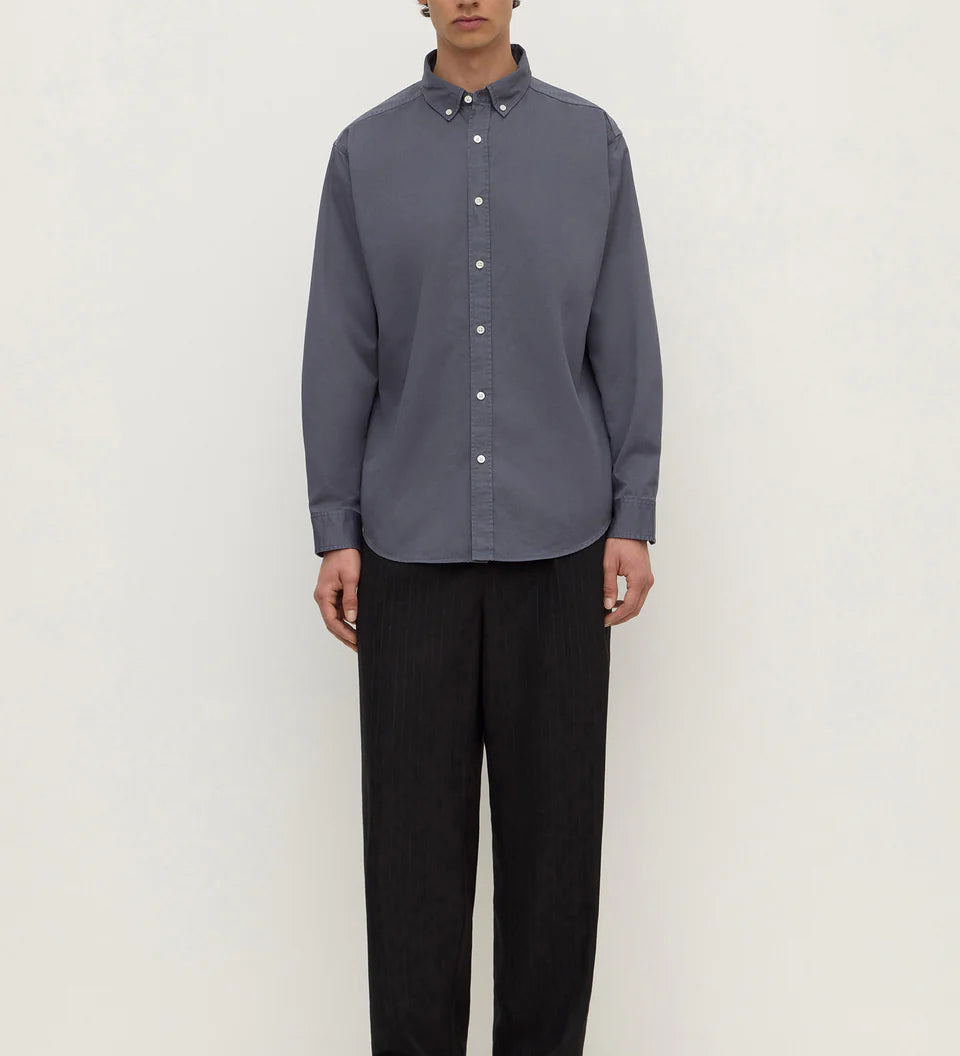 A man stands against a plain off-white studio background wearing a muted slate blue-gray long-sleeve button-down shirt with black trousers and black shoes.