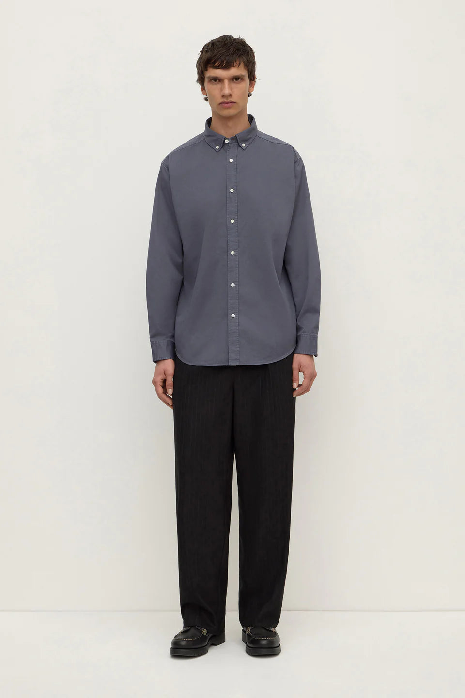 A man stands against a plain off-white studio background wearing a muted slate blue-gray long-sleeve button-down shirt with black trousers and black shoes.