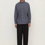 A man stands against a plain off-white studio background wearing a muted slate blue-gray long-sleeve button-down shirt with black trousers and black shoes.
