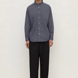 A man stands against a plain off-white studio background wearing a muted slate blue-gray long-sleeve button-down shirt with black trousers and black shoes.