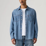 A man wearing a light blue denim button-up shirt over a white t-shirt and blue jeans, standing against a light gray studio background.