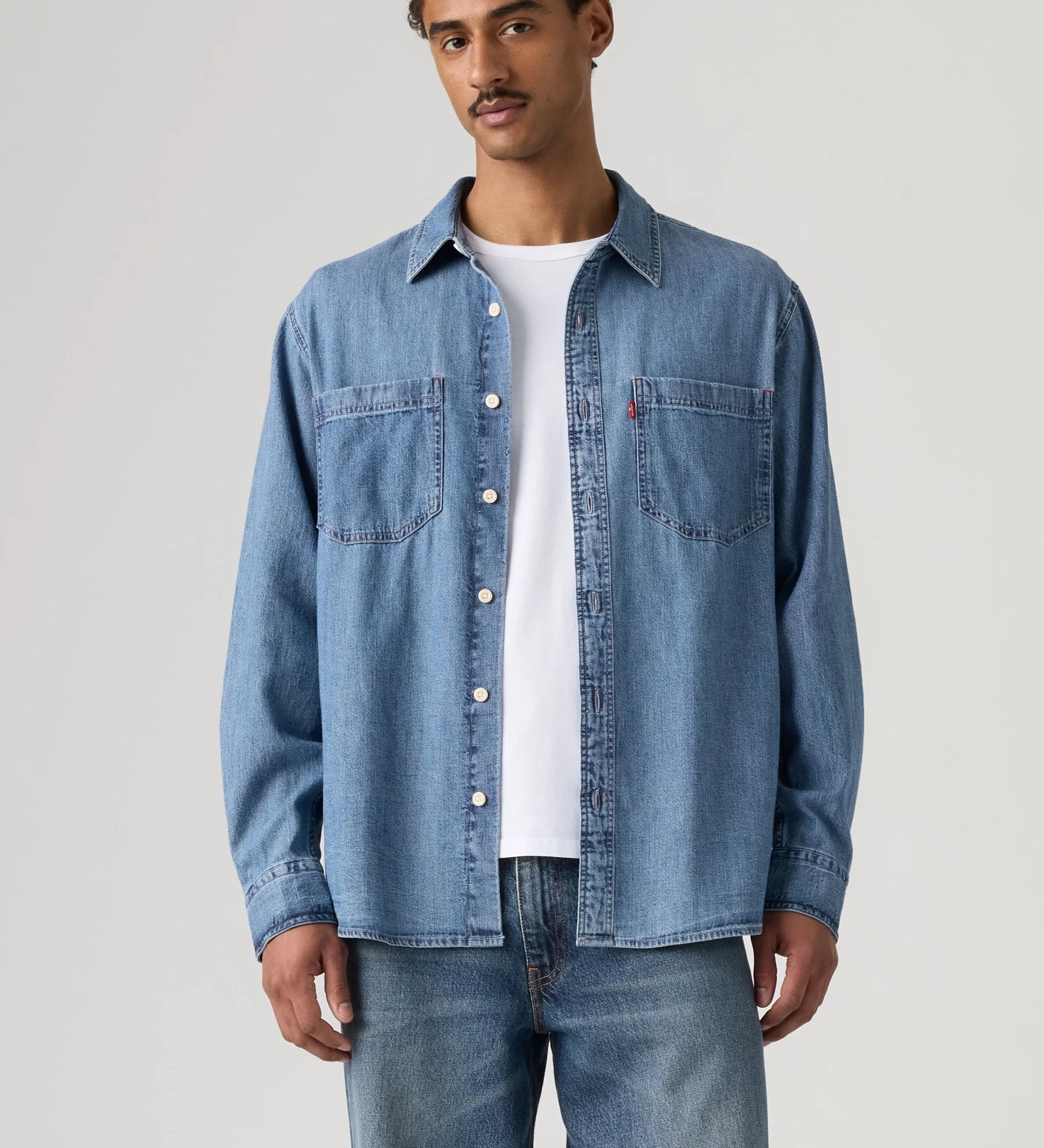 A man wearing a light blue denim button-up shirt over a white t-shirt and blue jeans, standing against a light gray studio background.