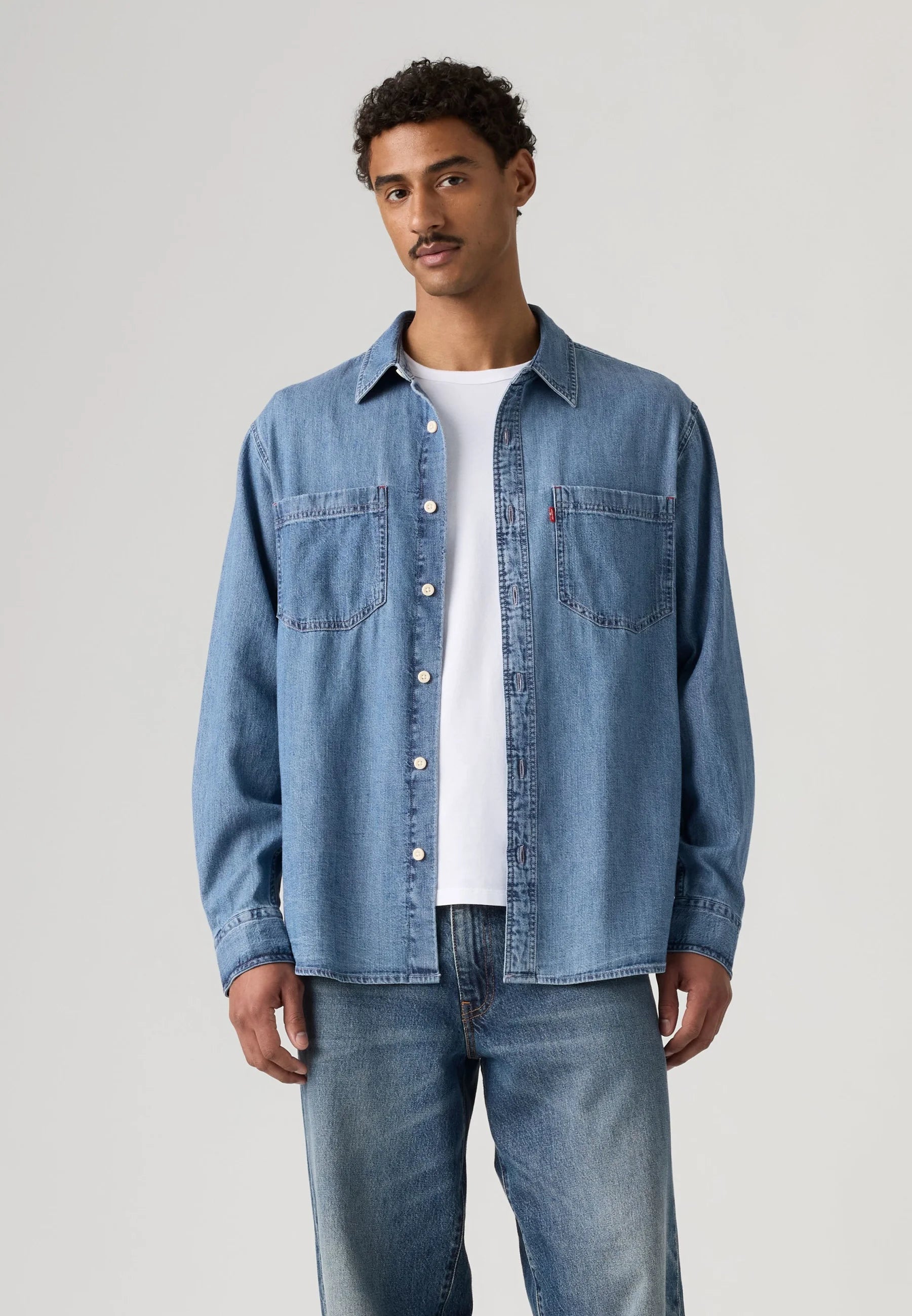 A man wearing a light blue denim button-up shirt over a white t-shirt and blue jeans, standing against a light gray studio background.
