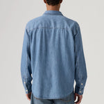 A man viewed from behind wearing a light blue denim long-sleeve button-up shirt and matching blue jeans, photographed against a neutral light gray studio background.