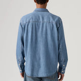 A man viewed from behind wearing a light blue denim long-sleeve button-up shirt and matching blue jeans, photographed against a neutral light gray studio background.