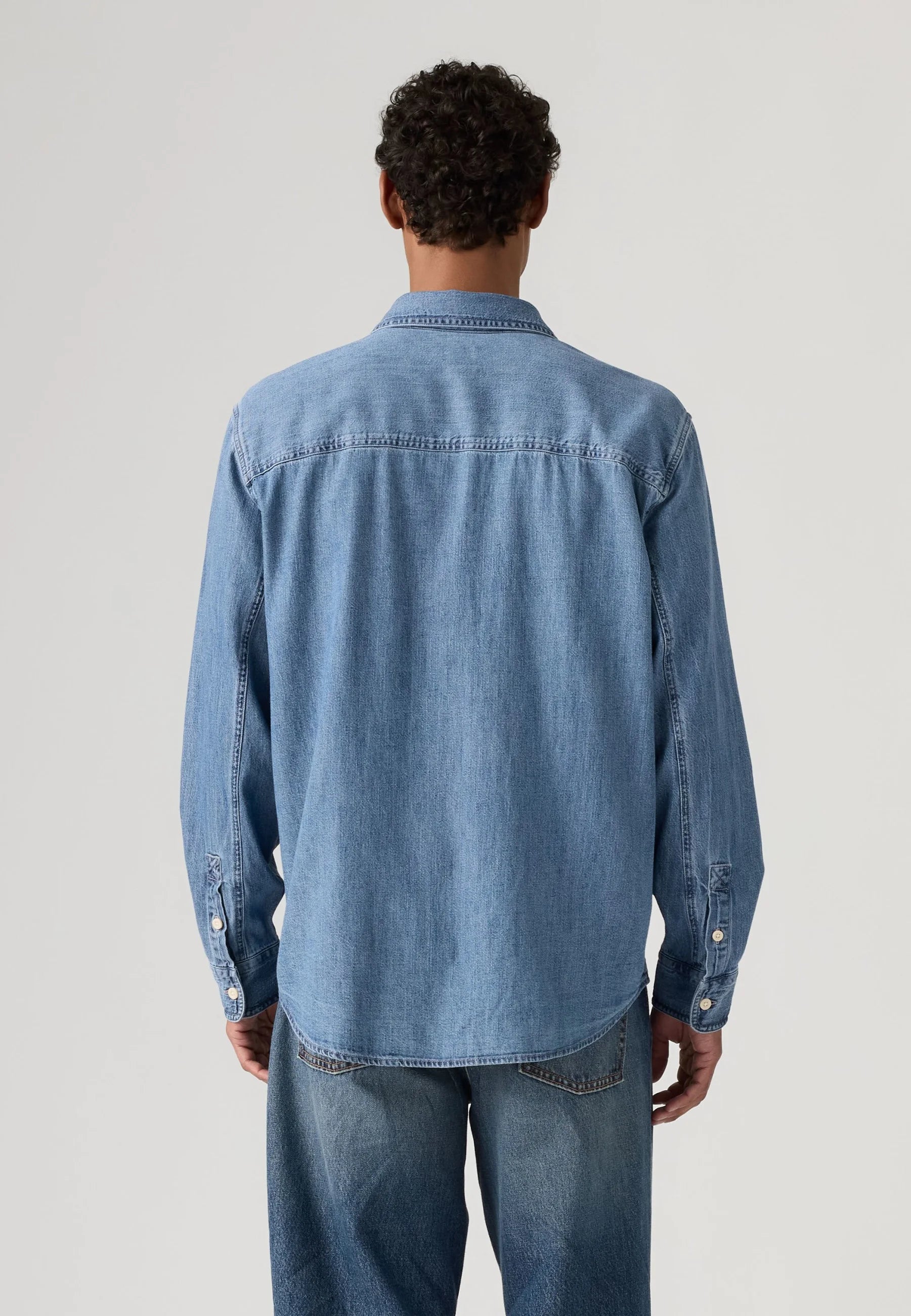 A man viewed from behind wearing a light blue denim long-sleeve button-up shirt and matching blue jeans, photographed against a neutral light gray studio background.