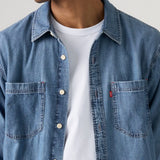 A close-up of a man wearing a light blue denim button-up shirt over a white t-shirt, showing the chest pocket with a small red Levi's tab on the left side.