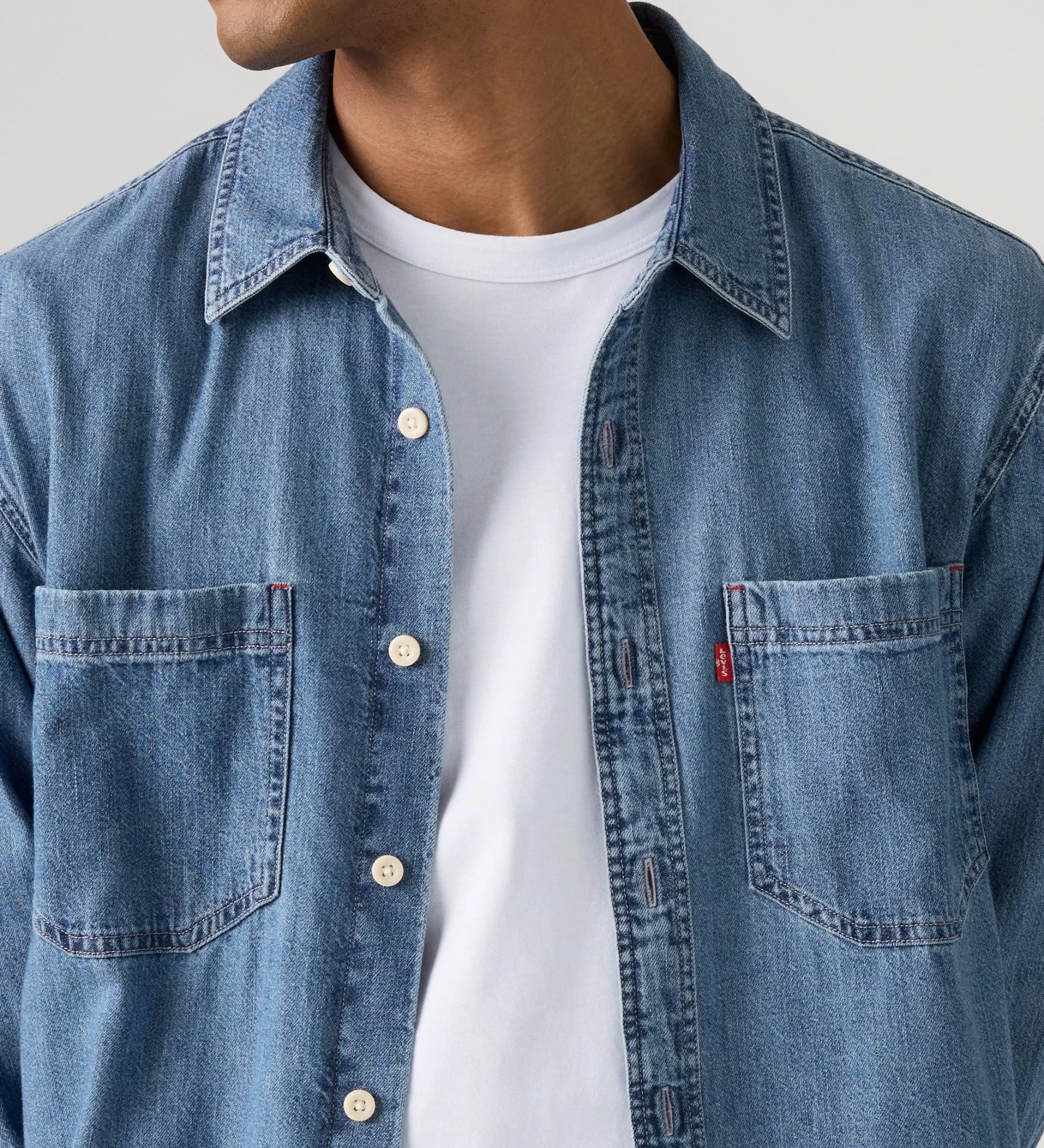 A close-up of a man wearing a light blue denim button-up shirt over a white t-shirt, showing the chest pocket with a small red Levi's tab on the left side.
