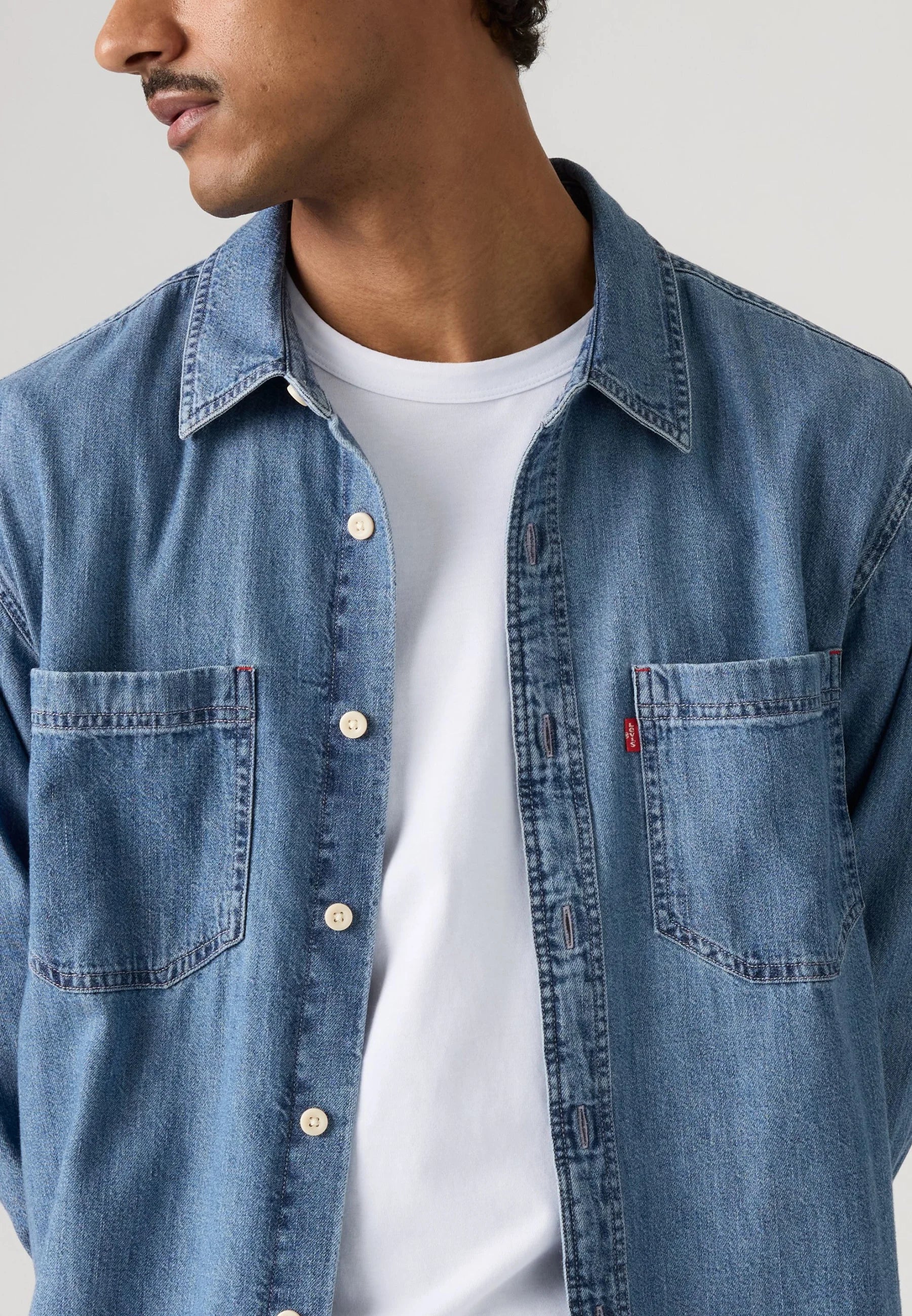 A close-up of a man wearing a light blue denim button-up shirt over a white t-shirt, showing the chest pocket with a small red Levi's tab on the left side.