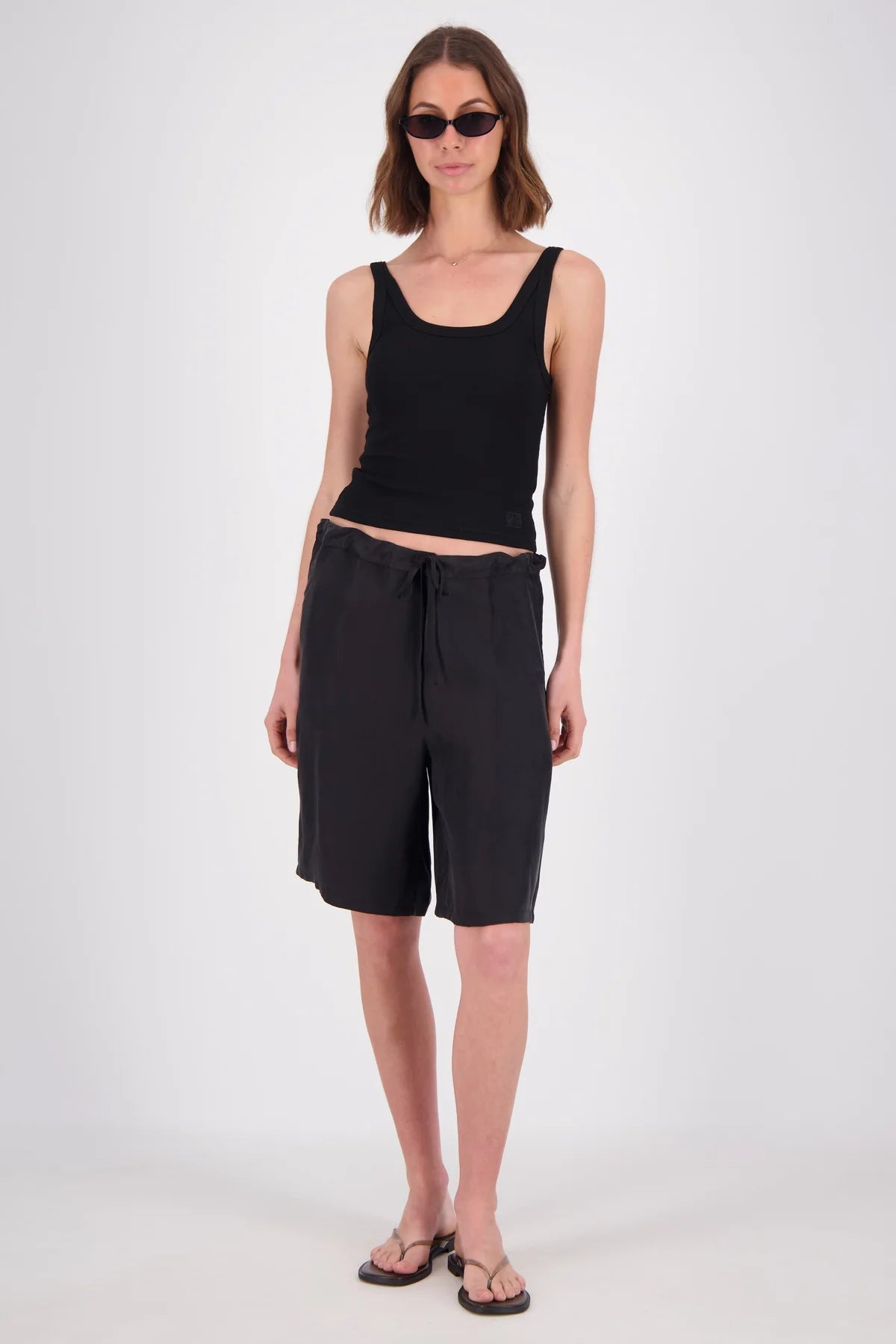 A model wearing a black ribbed tank top and black drawstring shorts, styled with black sunglasses and black sandals, standing against a light gray studio background.