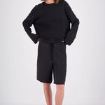 A woman modeling a black long-sleeve top and matching black shorts with a drawstring waist, styled with black sunglasses and dark sandals against a light gray studio background.
