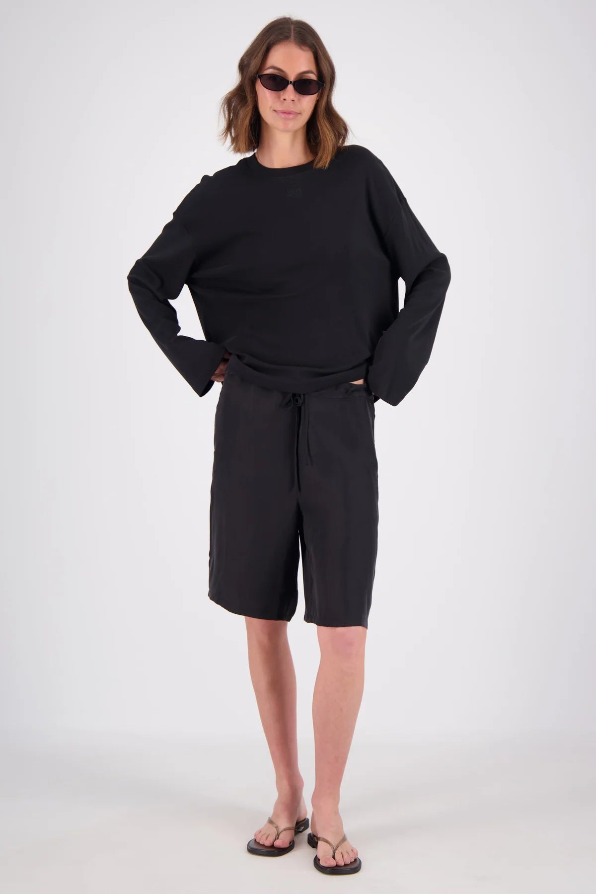 A woman modeling a black long-sleeve top and matching black shorts with a drawstring waist, styled with black sunglasses and dark sandals against a light gray studio background.