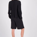 A model viewed from the back wearing a black long-sleeve top and matching black shorts, paired with black flip-flops, against a plain light gray studio background.