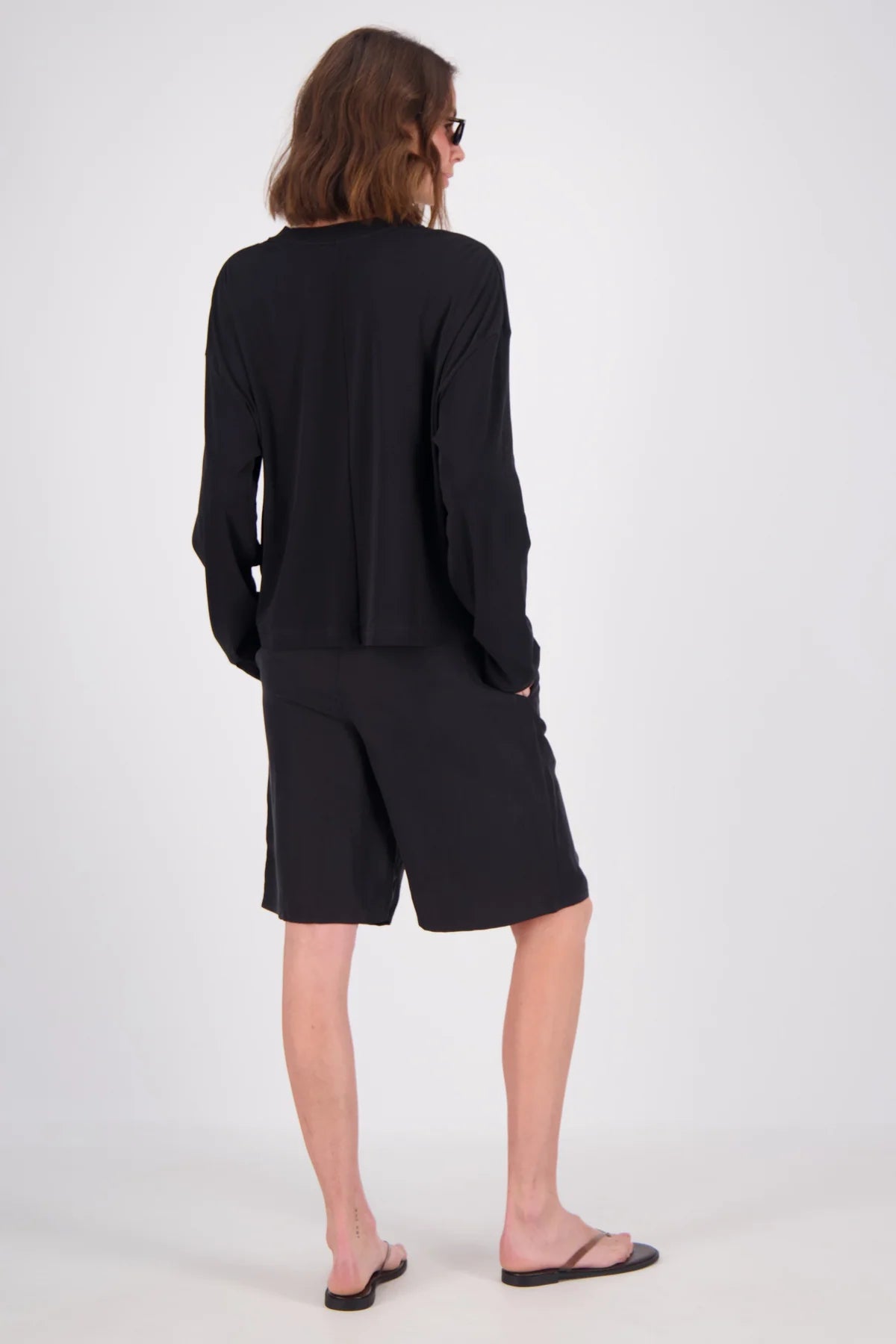 A model viewed from the back wearing a black long-sleeve top and matching black shorts, paired with black flip-flops, against a plain light gray studio background.