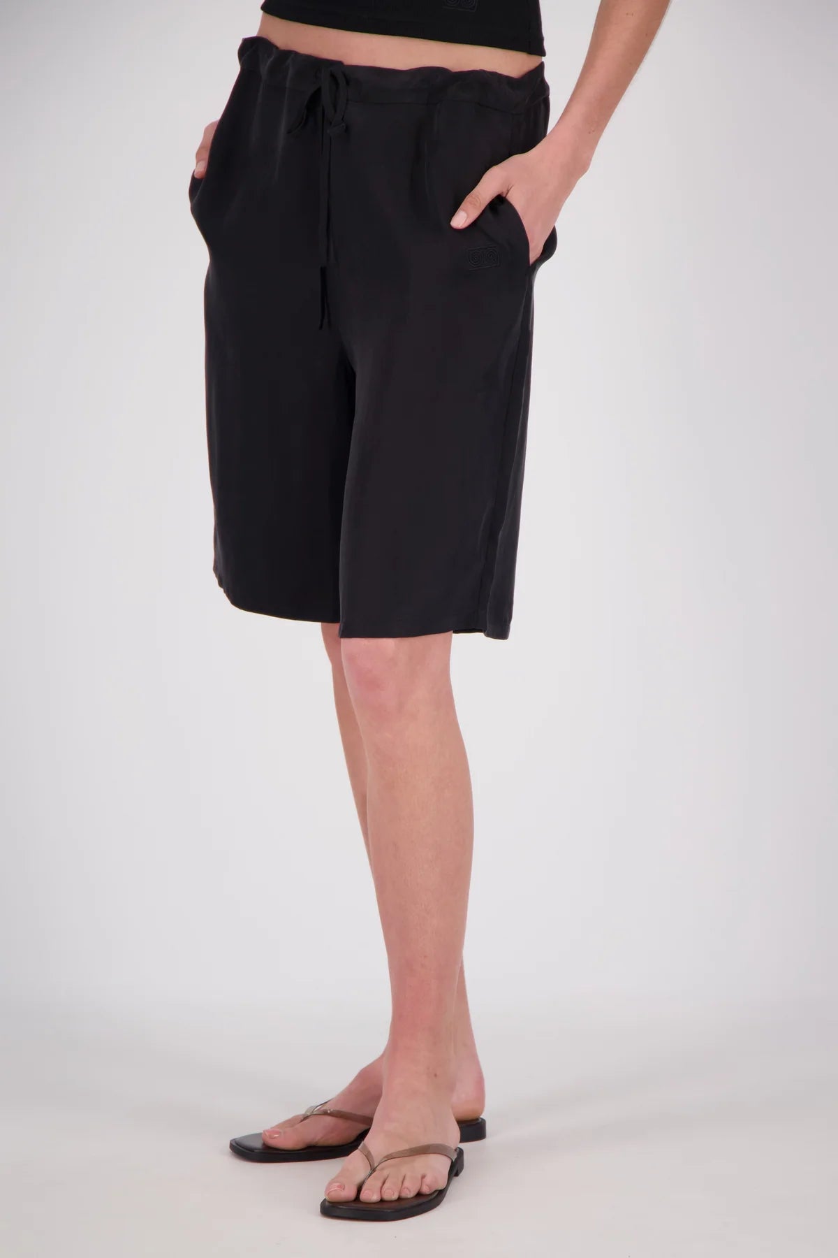 A person wearing black drawstring shorts with side pockets, paired with black sandals, shown from the waist down against a light gray studio background.