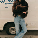 Person wearing a fur coat and jeans standing in front of a van with 'Warren' branding.