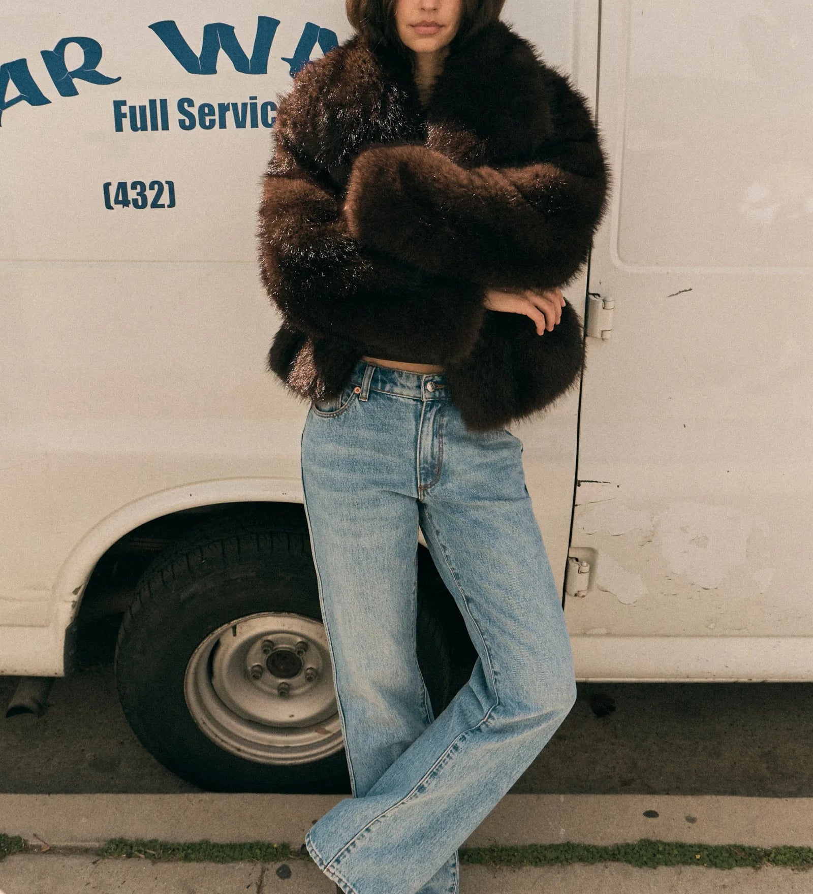 Person wearing a fur coat and jeans standing in front of a van with 'Warren' branding.