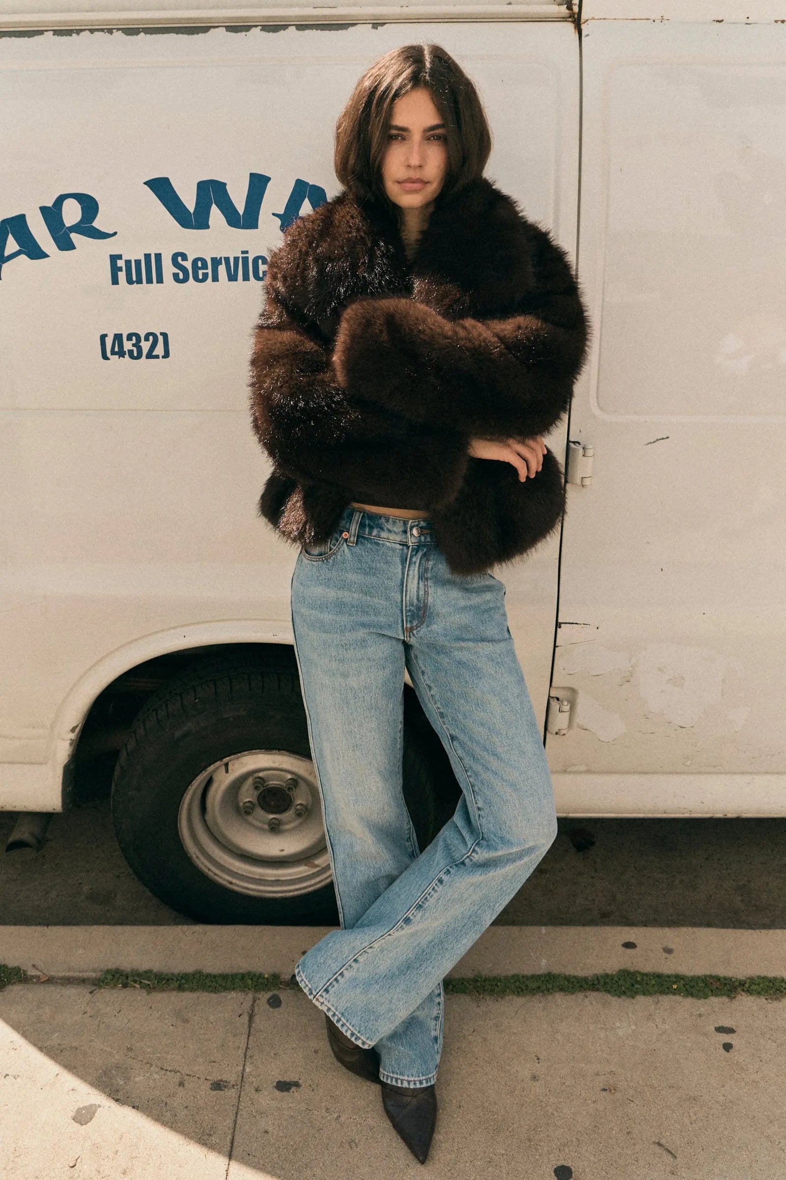 Person wearing a fur coat and jeans standing in front of a van with 'Warren' branding.