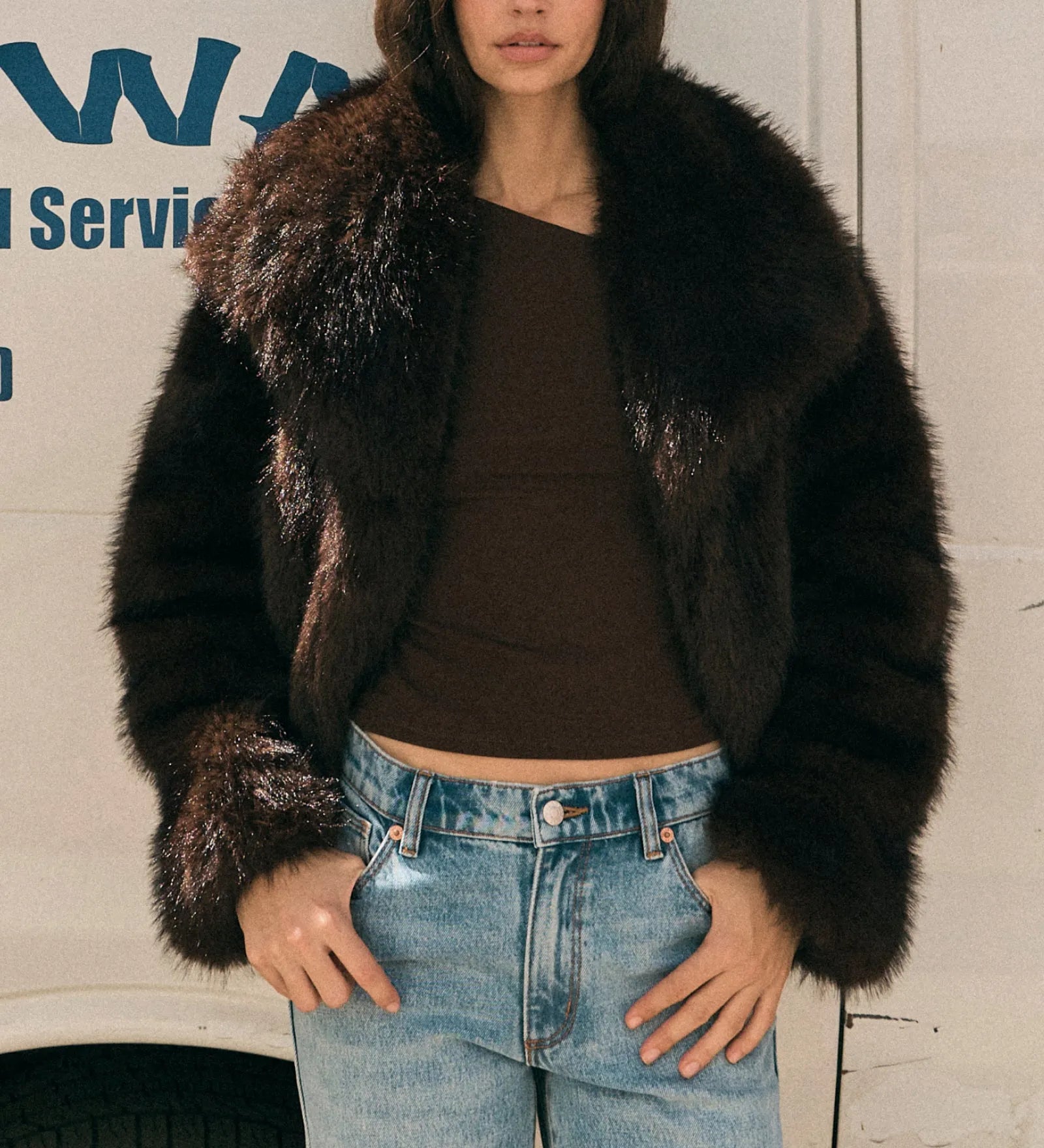 Woman wearing a fur coat and jeans standing in front of a white van with blue text.