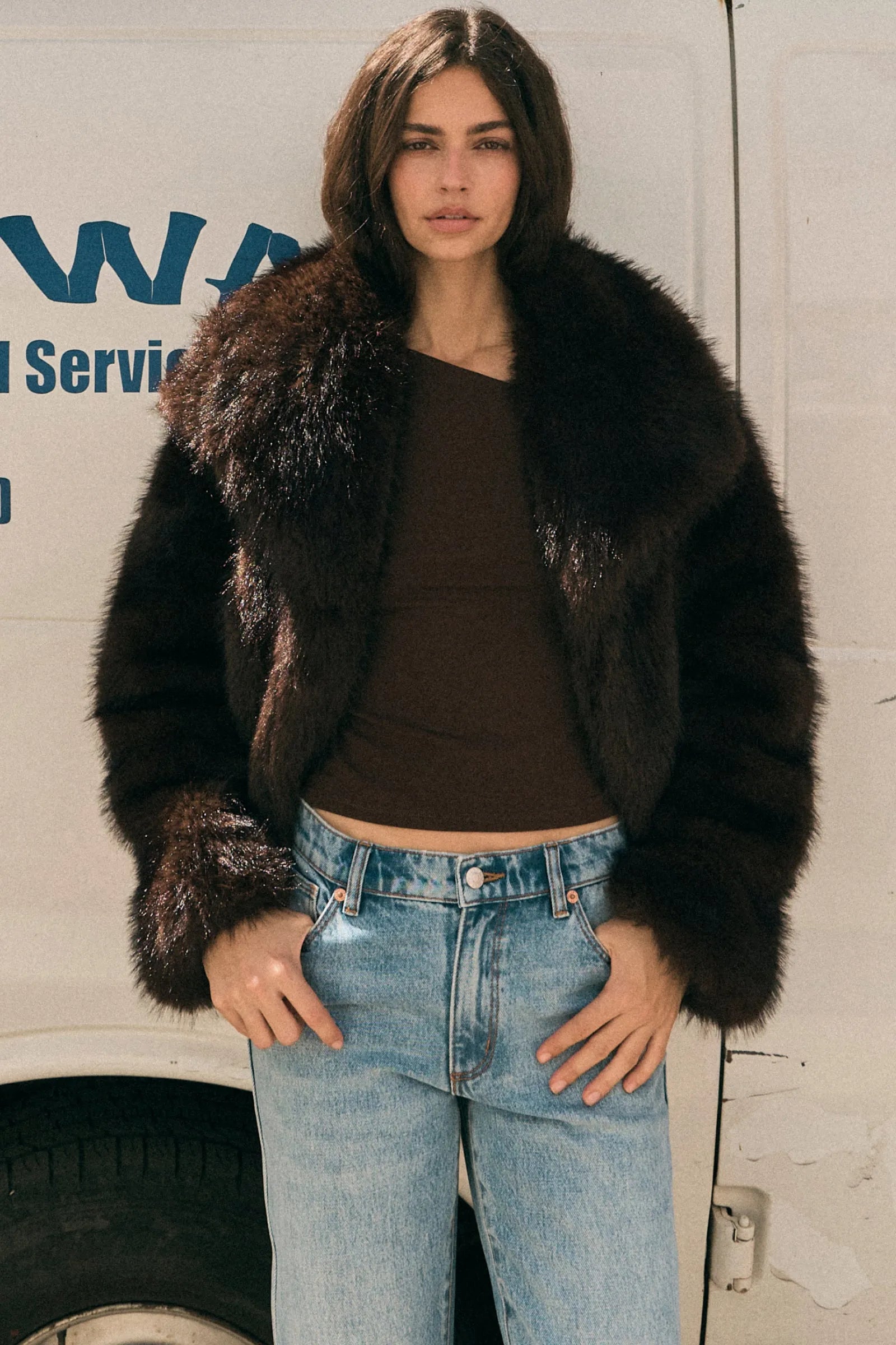 Woman wearing a fur coat and jeans standing in front of a white van with blue text.
