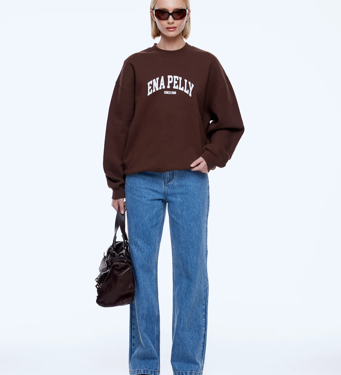 A model stands against a light gray studio background wearing a dark brown sweatshirt with 'ENA PELLY' printed in white across the chest, paired with blue jeans, black pointed shoes, sunglasses, and carrying a dark brown handbag.