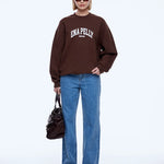 A model stands against a light gray studio background wearing a dark brown sweatshirt with 'ENA PELLY' printed in white across the chest, paired with blue jeans, black pointed shoes, sunglasses, and carrying a dark brown handbag.